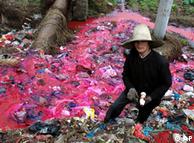 A woman collects plastic bottles near a river where water is polluted with a reddish dye 
