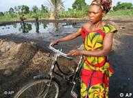 A Nigerian woman with a bicycle. In the background a leaking oil well