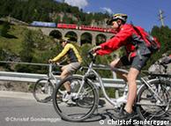 Молодые люди  едут на велосипедах на фоне гор и движущегося поезда